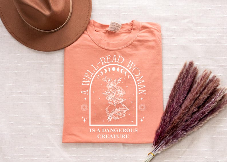 May include: Coral-colored t-shirt with the text "A WELL-READ WOMAN IS A DANGEROUS CREATURE" in white. The design includes an arched frame with a floral and book illustration. A brown hat and a bundle of dried flowers are also visible.