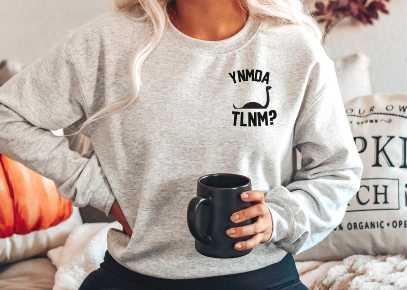 Puede incluir: Una sudadera gris con un gr&aacute;fico negro de un dinosaurio con el texto "YNMDA TLNM?" en la parte delantera. La sudadera la lleva puesta una persona que sostiene una taza de caf&eacute; negra.