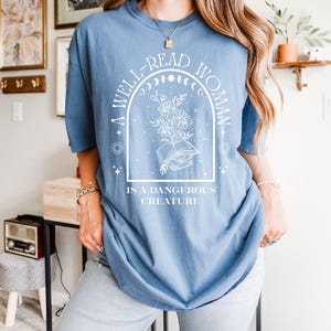 May include: A light blue t-shirt with the text "A WELL-READ WOMAN IS A DANGEROUS CREATURE" in a white graphic design. The design includes an open book, flowers, and celestial elements. The shirt is a casual, oversized fit.