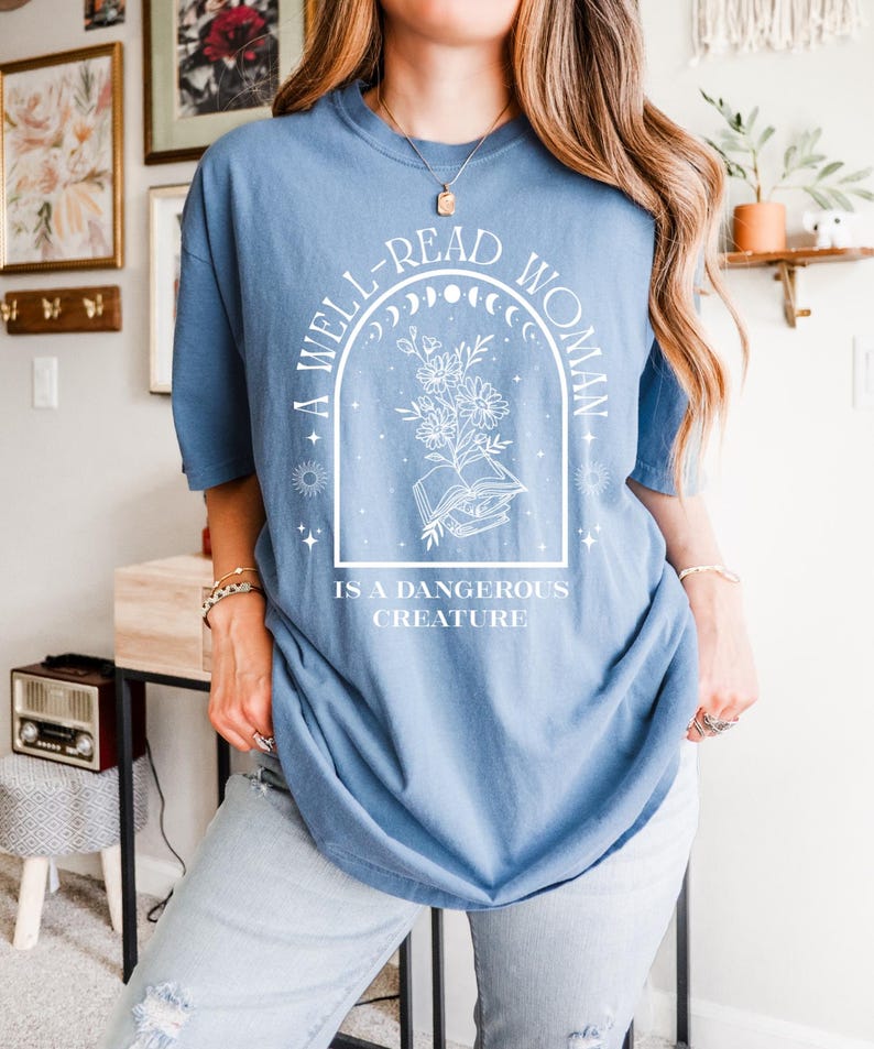 May include: A light blue t-shirt with the text "A WELL-READ WOMAN IS A DANGEROUS CREATURE" in a white graphic design. The design includes an open book, flowers, and celestial elements. The shirt is a casual, oversized fit.