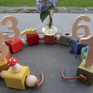 Könnte beinhalten: Ein hölzerner Spielzeugzug mit bunten Blöcken, die die Zahlen 1 bis 8 zeigen. Jeder Block hat eine andere Farbe und ein anderes Design, darunter ein Blitz, ein Pilz und ein Marienkäfer. Der Zug ist durch eine braune Lederschnur verbunden.