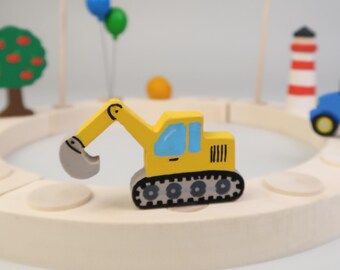 May include: A yellow toy excavator with a black scoop and black tracks is on a wooden circular track with a blue toy truck, a red and white lighthouse, and a green tree with red apples.