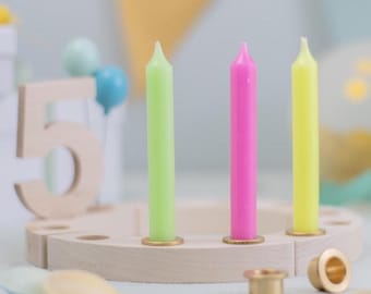 May include: A wooden birthday candle holder with three colorful candles: green, pink, and yellow. The candles are set in a light-colored wooden base with gold-colored metal holders. The number 5 is visible in the background, along with a cake.