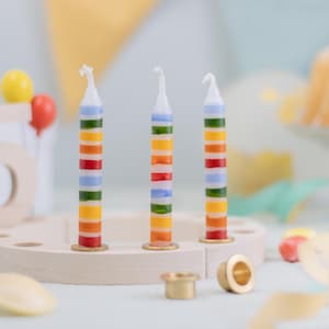 May include: Three colorful striped birthday candles on a wooden candle holder. The candles have red, orange, yellow, green, and blue stripes. A wooden number 5 and balloons are in the background.