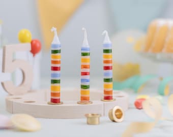 May include: Three colorful striped birthday candles on a wooden candle holder. The candles have red, orange, yellow, green, and blue stripes. A wooden number 5 and balloons are in the background.