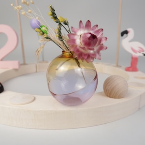 Könnte beinhalten: Eine kleine, runde, klare Glasvase mit einem rosa-gelben Ombre-Effekt. Die Vase ist mit getrockneten Blumen gefüllt, darunter eine rosa Blume mit gelber Mitte.