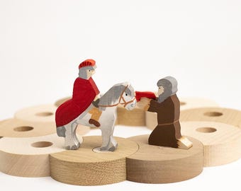 May include: A wooden toy set featuring a figure in a red cloak riding a gray horse and a kneeling figure in a brown robe, both positioned on a circular base made of several light-colored wooden rings with central holes.