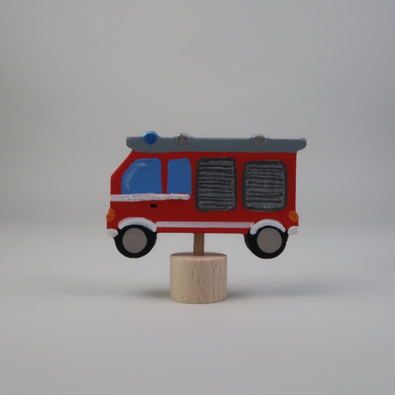 May include: A red and white toy fire truck with a blue light on top, on a wooden base. The truck has black wheels and a white stripe around the middle.