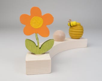 May include: A wooden toy set with a bee, a honeycomb, a flower, and a ball. The bee is yellow and black, the honeycomb is yellow, the flower is orange with a yellow center, and the ball is brown.