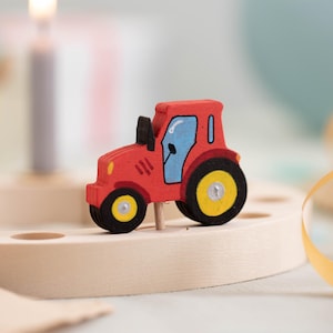 Könnte beinhalten: Ein roter Holztrecker mit schwarzen Rädern und einer blauen Kabine. Der Spielzeugtrecker hat gelbe Akzente und ist auf einem Holzsockel montiert. Das Bild ist eine Nahaufnahme mit unscharfem Hintergrund.