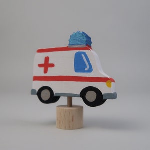 May include: A white toy ambulance with a red cross, blue light, and yellow details. The ambulance is on a wooden base.
