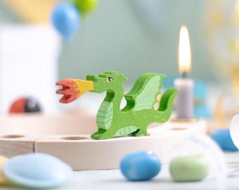 May include: A green wooden dragon figurine with red and yellow flames emanating from its mouth. The dragon is perched on a light-colored wooden base, with a lit candle and colorful decorations in the background. A whimsical, decorative item.