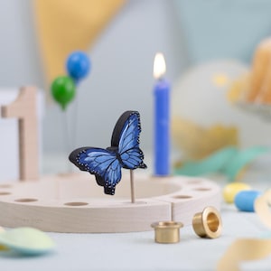 Könnte beinhalten: Ein hölzerner Geburtstagskuchenständer mit einer blauen Schmetterlingsdekoration, einer blauen Kerze und der Zahl 1. Der Schmetterling hat blaue Flügel mit schwarzen Rändern und weißen Punkten. Zwei kleine Ballons sind im Hintergrund.