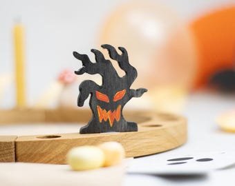 May include: A black wooden cutout of a gnarled, spooky tree with a jagged, orange-painted face featuring sharp eyes and a wide, toothy grin, standing upright on a curved, light-colored wooden base with circular indentations.