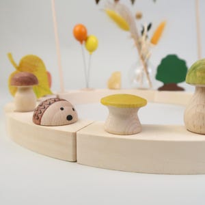Könnte beinhalten: Ein Holzspielzeugset mit einer kreisförmigen Bahn, bestehend aus einem Igel, Pilzen und einem Blatt. Die Bahn ist aus hellem Holz gefertigt und die Figuren sind in natürlichen Farben bemalt.