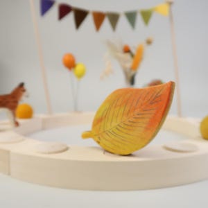 Könnte beinhalten: Ein Holzspielzeugblatt in Gelb- und Orangetönen mit detaillierten Adern. Das Blatt ist Teil eines kreisförmigen Holzspielsets mit einer Fuchsfigur, Luftballons und einem Banner im Hintergrund. Die Szene erinnert an den Herbst.
