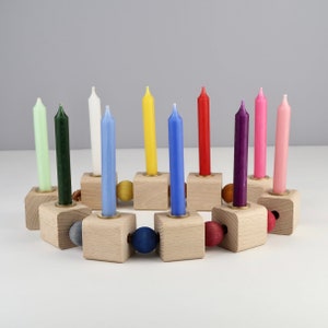 Könnte beinhalten: Ein Satz mit 10 Kerzenhaltern aus Holz mit bunten Kerzen. Die Halter sind kreisförmig miteinander verbunden und haben jeweils eine kleine Kugel dazwischen. Die Kerzen sind in verschiedenen Farben, darunter Rot, Blau, Gelb, Grün und Pink.