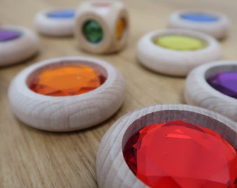 May include: A collection of light-colored wooden rings and a wooden cube, each featuring inset faceted acrylic gems in various colors including red, orange, yellow, green, blue, and purple. The smooth wooden surfaces contrast with the translucent gems.