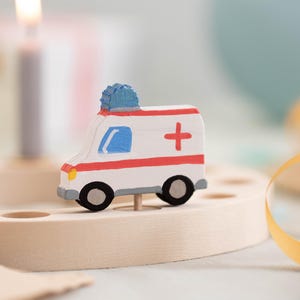 May include: A white toy ambulance with a red cross and blue light on top. The ambulance is on a wooden base.