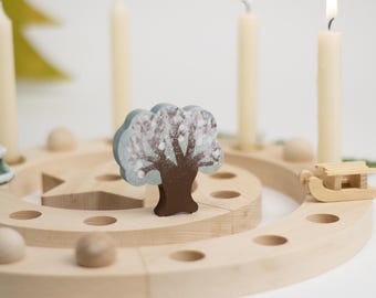May include: A circular wooden advent ring featuring multiple holes for candles, decorated with a small painted tree, a miniature wooden sled, a green tree figurine, and four tall cream-colored taper candles standing upright.