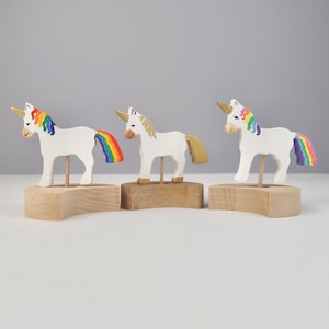 May include: Three wooden unicorn figurines with rainbow manes and tails. Each unicorn is standing on a wooden base.