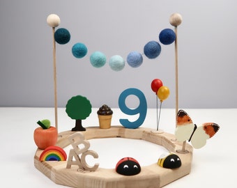 May include: A wooden cake topper with a blue number nine, a blue and white felt garland, a red apple, a green tree, a cupcake, a rainbow, the letters B, A, and C, a ladybug, a bee, and a butterfly.