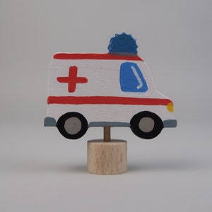 May include: A wooden toy ambulance with red and white stripes, a blue light on top, and a red cross painted on the side. The ambulance is on a wooden base.