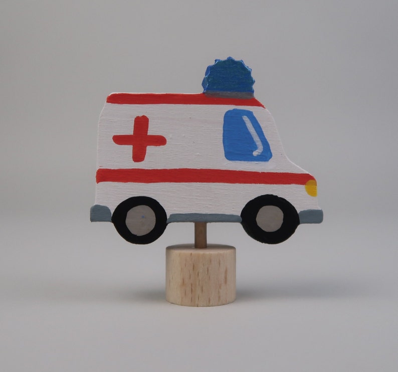 May include: A wooden toy ambulance with red and white stripes, a blue light on top, and a red cross painted on the side. The ambulance is on a wooden base.