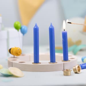 May include: A wooden candle holder with three blue candles, one lit by a match. The holder has brass accents and is surrounded by decorative items, including a bee figurine and balloons. The background includes gift boxes and a cake.