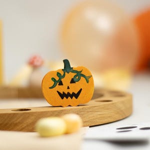Könnte beinhalten: Ein kleiner, flacher Kürbis-Ausschnitt aus orangefarbenem Holz mit einem handgemalten schwarzen Jack-o'-Lantern-Gesicht und dunkelgrünen Rankendetails am Stiel, aufrecht stehend auf einem geschwungenen, hellen Holzsockel mit kreisförmigen Vertiefungen.