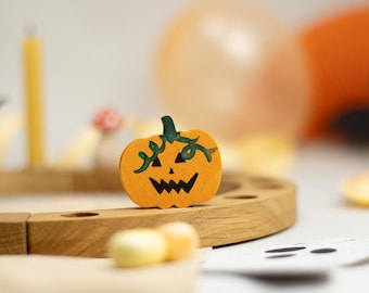May include: A small, flat, orange wooden pumpkin cutout featuring a hand-painted black jack-o'-lantern face and dark green vine details on the stem, positioned upright on a curved, light-colored wooden base with circular indentations.