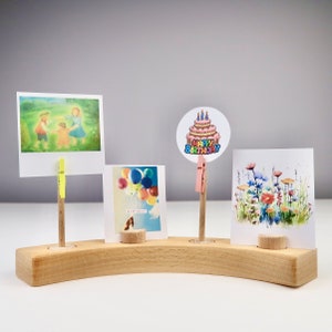 Könnte beinhalten: Ein Holzständer mit drei Schlitzen zum Ausstellen von Karten oder Fotos. Der Ständer ist gebogen und hat eine natürliche Holzoptik. Auf dem Ständer sind drei Karten ausgestellt, eine mit einem Geburtstagskuchen, eine mit Ballons und eine mit einem Bild von Kindern.