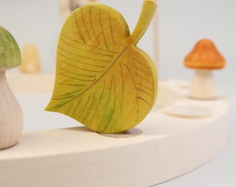 May include: A wooden toy with a yellow leaf, a green mushroom, and an orange mushroom. The toy is made of natural wood and is painted with non-toxic paints.