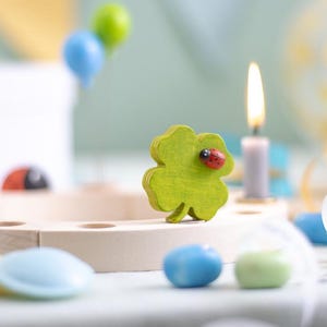 Könnte beinhalten: Eine hölzerne Kleeblatt-Dekoration mit einem roten Marienkäfer oben, umgeben von bunten Süßigkeiten und einer weißen Kerze auf einem Holzsockel.