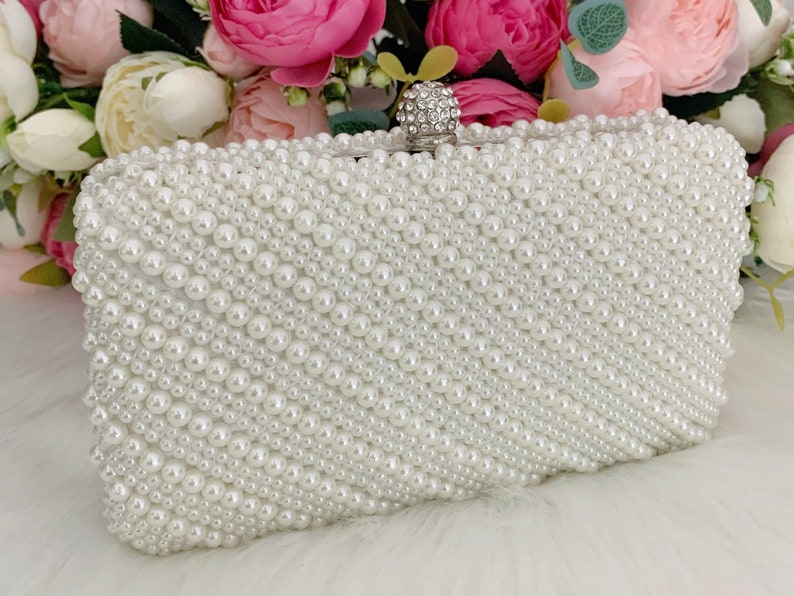 Peut inclure: Une pochette blanche recouverte de fausses perles. La pochette a un fermoir argent&eacute; avec un embellissement en strass transparent.