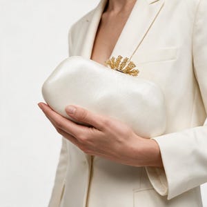 May include: A cream-coloured satin clutch bag with a smooth, slightly textured finish. The bag features a gold-toned metal clasp at the top, designed with an intricate, fan-like structure that resembles a cluster of small, textured metallic petals.
