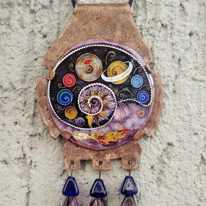 Op de afbeelding: Een koperen ketting met een ronde hanger van zwart emaille met een spiraal, planeten en sterren. De hanger is bevestigd aan een bruin leren koord en heeft drie blauwe en paarse glazen kralen die eraan hangen.