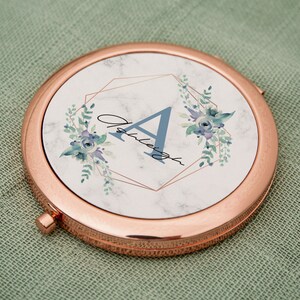 Puede incluir: Un espejo compacto de oro rosa con un diseño de mármol blanco y un patrón floral azul. El espejo tiene un marco geométrico dorado y la letra "A" en el centro.