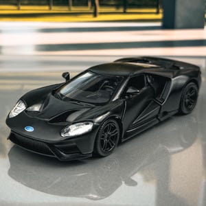 Può includere: Un'auto modello nera, una Ford GT, è esposta su una superficie riflettente. Il veicolo in miniatura presenta dettagli, tra cui fari, una griglia anteriore e il logo Ford. Lo sfondo mostra una scena esterna sfocata con alberi.