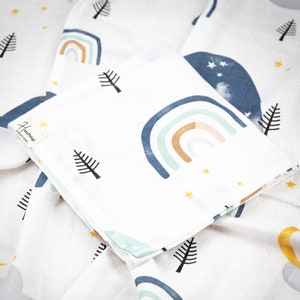 May include: A white muslin swaddle blanket with a rainbow, stars, and trees pattern. The blanket is folded in half and has a small tag that says "Hummingbird Creations".