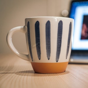 Puede incluir: Una taza de cerámica blanca con rayas verticales azules y un fondo naranja. La taza está sobre una mesa de madera delante de un portátil.