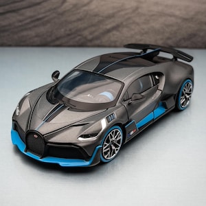 May include: A gray and blue model sports car with black accents. The car has a sleek design with blue trim on the front, sides, and wheels. A black spoiler is visible on the back of the car. The background shows a landscape with mountains.
