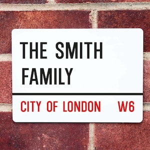May include: White sign with black text that reads "THE SMITH FAMILY CITY OF LONDON W6". The sign is mounted on a red brick wall.