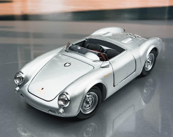 Può includere: Modellino d'auto argentata con interni neri. Il veicolo in miniatura ha un design roadster classico con un profilo basso e una capote aperta. L'auto è ambientata su uno sfondo sfocato di alberi ed elementi architettonici.