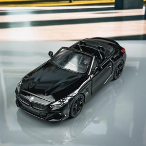 Könnte beinhalten: Ein glänzend schwarzes Miniatur-Cabriolet, ein BMW Z4, mit detailliertem Frontgrill und schwarzen Rädern. Das Spielzeugauto steht auf einer reflektierenden Oberfläche. Der Hintergrund zeigt eine verschwommene Außenszene mit Bäumen.