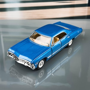 Op de afbeelding: Een blauwe modelauto met whitewall-banden en een chromen grille. De klassieke auto heeft een beige interieur en staat tegen een vage achtergrond van bomen en architectuur.