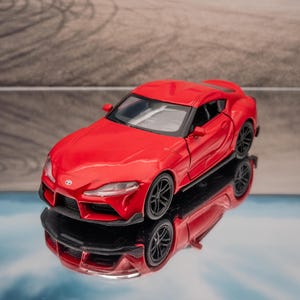 May include: A red Toyota Supra die-cast model car featuring a sleek aerodynamic body, black multi-spoke wheels, and clear windows. The miniature vehicle is positioned on a reflective surface, showcasing its detailed front grille and headlights.