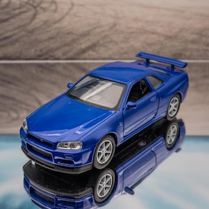 Könnte beinhalten: Ein blaues Modellauto des Sportwagens Nissan Skyline GT-R R34 mit Heckspoiler, silbernen Mehrspeichenfelgen, klaren Scheinwerfern und einem schwarzen Frontgrill, dargestellt aus der Vogelperspektive mit einer deutlichen Spiegelung.