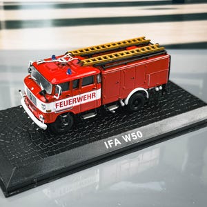 May include: A red IFA W50 fire engine model with a yellow ladder on top. The model has the word "FEUERWEHR" in white on the side. The model is on a black base with the text "IFA W50".