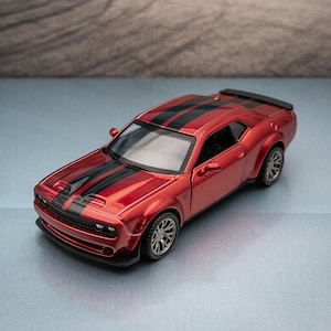 Puede incluir: Un coche de juguete rojo con rayas negras en el capó y el techo. El coche tiene ruedas plateadas y un alerón negro. El fondo presenta un paisaje borroso con montañas y un lecho de lago seco.